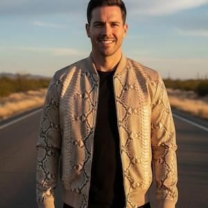 Może przedstawiać: Beżowa kurtka bomberka ze wzorem wężowej skóry. Kurtka ma zapięcie na zamek błyskawiczny oraz prążkowane mankiety i dół. Osoba ma na sobie czarny t-shirt. Tło to droga z błękitnym niebem.