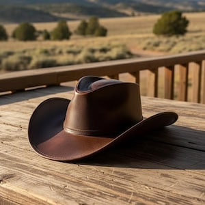 May include: A brown leather cowboy hat with a wide brim rests on a weathered wooden table. The hat has a classic design with a slightly curved crown and a band around the base. The background shows a rural landscape.