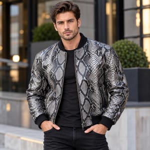 Può includere: Un giubbotto bomber con motivo a pelle di serpente argento e nero, dotato di colletto a coste nero, polsini a coste neri e una cerniera centrale in metallo. La giacca è indossata sopra una t-shirt nera semplice e abbinata a jeans neri.