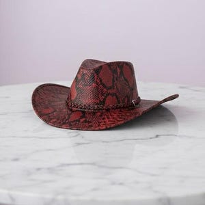 May include: A red cowboy hat with a snakeskin pattern. The hat has a wide brim and a decorative band around the crown. The hat is resting on a white marble table.