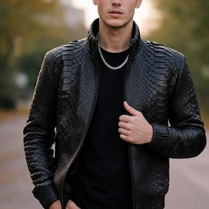 Może przedstawiać: Czarna skórzana kurtka z teksturowanym wzorem przypominającym skórę gada. Kurtka ma zamek błyskawiczny z przodu, stójkę i prążkowane mankiety. Osoba ma na sobie czarny t-shirt, srebrny łańcuszek i niebieskie dżinsy.