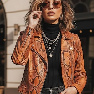 Pode incluir: Casaco de couro laranja com padrão de pele de cobra, gola alta preta, colares em camadas e óculos de sol redondos. O casaco tem fecho de correr e um design clássico de casaco de motociclista. O conjunto é completado com jeans pretos.