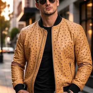Può includere: Giacca bomber in pelle color cuoio con motivo a pois traforati. La giacca ha colletto, polsini e chiusura con cerniera neri. La persona indossa una maglietta nera, occhiali da sole e un orologio.