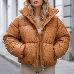 Könnte beinhalten: Eine hellbraune Daunenjacke mit texturiertem Straußenleder-Muster, ausgestattet mit einem hohen Kragen, langen Ärmeln und einem Frontreißverschluss. Die Jacke hat ein gestepptes Design mit horizontalen Nähten und zwei seitlichen Eingrifftaschen.