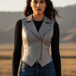 以下が含まれることがあります： ジッパー開閉、ラペルカラー、スタッズディテールが特徴のライトグレーのレザーベスト。黒の長袖シャツの上に着用し、ブルージーンズと合わせています。ベストはテクスチャード加工されたドット柄です。