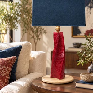 May include: A table lamp featuring a rectangular navy blue fabric shade and a twisted, textured red base with a crocodile-skin pattern. The lamp stands on a circular light-colored stone base and includes a brass-colored metal socket.