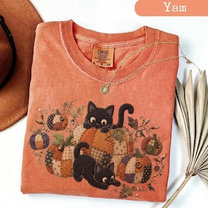 Puede incluir: Una camiseta de cuello redondo color naranja quemado con un gráfico de dos gatos negros y varias calabazas estilo patchwork. La camiseta tiene una etiqueta Comfort Colors en el cuello interior. Una fina cadena dorada descansa sobre la tela.