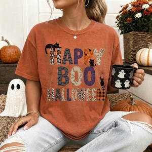 Puede incluir: Una camiseta de manga corta color naranja quemado con el texto Happy BOO Halloween en letras de bloque estampadas. El diseño incluye ilustraciones de gatos negros, fantasmas, calabazas y huesos. La camiseta se acompaña de una taza negra con fantasmas blancos.