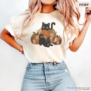 Puede incluir: Una camiseta de color marfil de manga corta con un estampado gráfico de dos gatos negros sentados entre tres calabazas estampadas. Las calabazas muestran un diseño de patchwork en tonos naranja, marrón y beige. El texto IVORY está arriba a la derecha.
