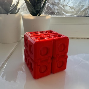 May include: A vibrant red geometric cube made up of smaller, connected blocks. Each block has a circular aperture and a rectangular design. The cube is on a white surface, with potted plants in the background.