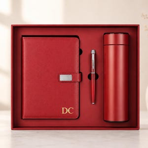 Könnte beinhalten: Ein rotes Geschenkset bestehend aus einem Notizbuch mit silberfarbenem Metallverschluss und goldgeprägten Buchstaben DC, einem passenden roten Kugelschreiber mit silbernen Akzenten und einer zylindrischen roten Isolierflasche in einer roten Box.