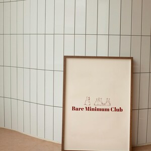 Puede incluir: Un cuadro enmarcado con fondo beige claro y el texto "Bare Minimum Club" en rojo oscuro. El cuadro presenta tres pequeñas ilustraciones de animales. El marco es de color marrón oscuro.