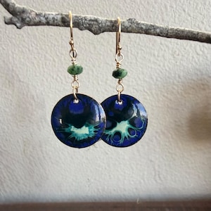 May include: A pair of dangle earrings featuring circular, enameled metal discs with a vibrant blue, black, and white abstract pattern. Each earring is suspended from a gold-toned wire hook by a small, faceted green gemstone bead and wire.