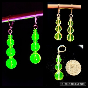 Pode incluir: Um par de brincos de contas de vidro verde com ganchos de prata. Os brincos têm três contas cada um, e as contas são redondas e translúcidas.