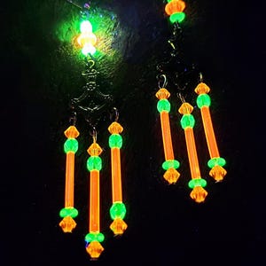 Pode incluir: Um par de brincos pendentes com contas laranja e verdes que brilham no escuro. Os brincos têm uma base de metal preto e são adornados com pequenas contas transparentes.