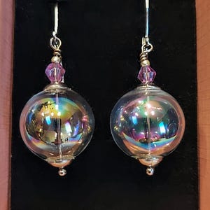 May include: A pair of iridescent glass sphere earrings with pink crystal accents. The earrings are silver-toned and have a dangling design.