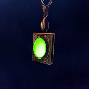 May include: A bronze-colored pendant necklace featuring a rectangular charm with an oval, glowing green stone. The charm has an ornate, vintage design. The necklace chain is also bronze-colored, creating a cohesive, antique aesthetic.