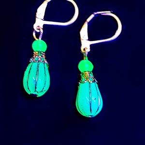 Pode incluir: Um par de brincos de gota de vidro verde com detalhes dourados e laços de arame.
