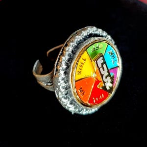 Könnte beinhalten: Ein silberner Ring mit einem bunten Raddesign. Das Rad hat Segmente in Gelb, Orange, Rot und Grün, mit den Wörtern "THIS", "ME", "THAT" und "OFF". In der Mitte steht das Wort "FUCK".
