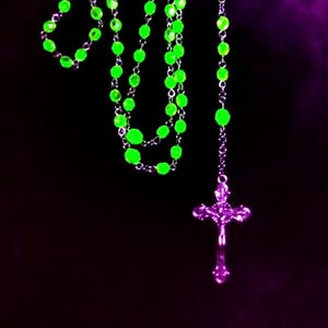 May include: A green beaded rosary with a silver cross pendant. The beads are glowing in the dark, and the cross is reflecting a purple light.