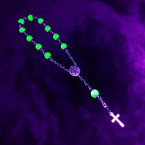 May include: A rosary with glowing green beads and a silver cross pendant. The beads are connected by a silver chain, and the background is a dark purple color.