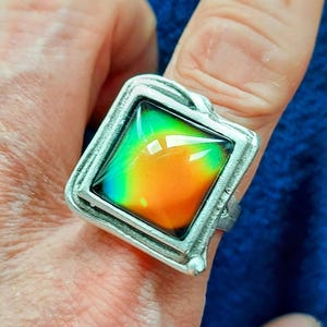 May include: A silver-toned ring with a square, color-changing stone. The stone transitions from green to orange, creating a vibrant, eye-catching effect. The ring has a textured, ornate setting, adding to its unique design. A piece of jewelry.