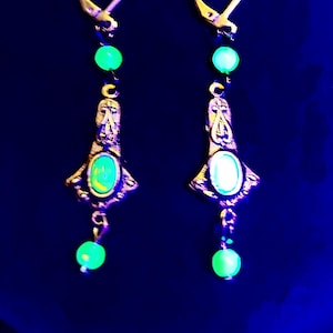 Pode incluir: Um par de brincos pendentes com contas de vidro verde e um design de metal dourado. Os brincos apresentam uma pedra verde em forma de lágrima.
