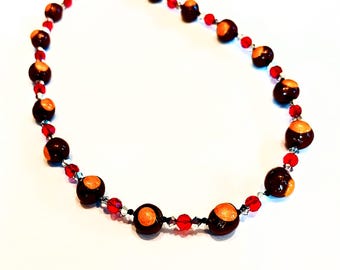 May include: A necklace featuring dark brown and orange beads, interspersed with small red beads. The necklace has a black clasp and is set against a white background. The beads appear to be made of a natural material.