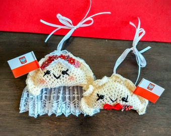May include: Two crocheted pierogi ornaments, one wearing a white veil and holding a Polish flag, the other wearing a red bow tie and holding a Polish flag.
