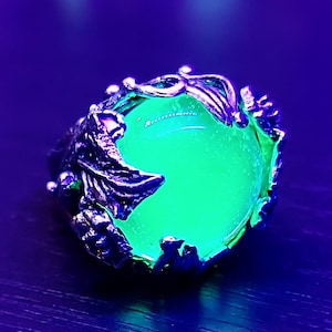 May include: A silver ring with a large, round, green gemstone set in a detailed, leafy design. The stone glows brightly under a black light.