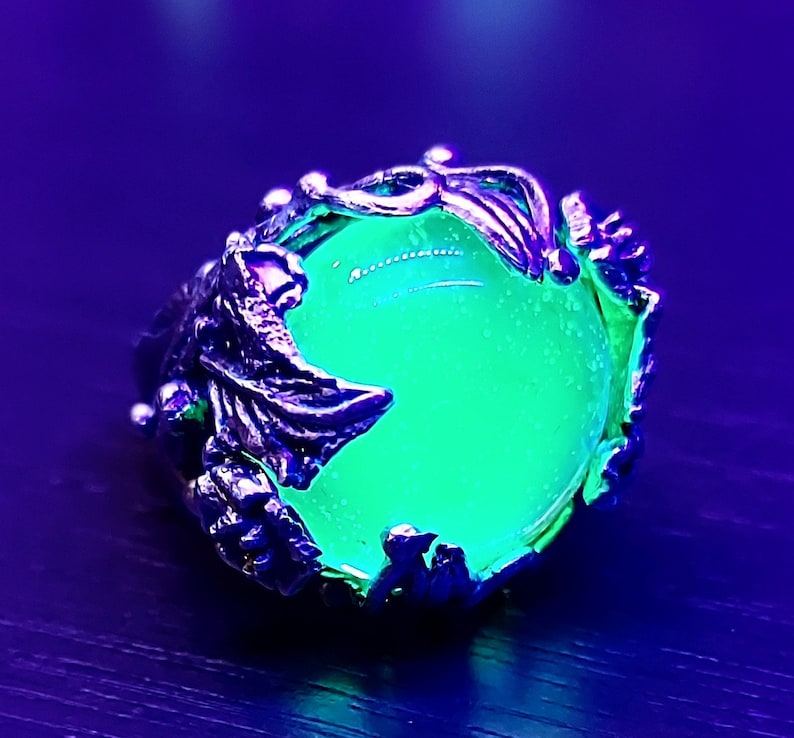 May include: A silver ring with a large, round, green gemstone set in a detailed, leafy design. The stone glows brightly under a black light.