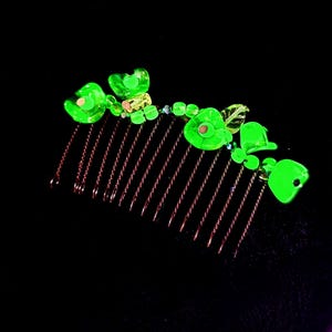 May include: A hair comb featuring copper-coloured teeth and vibrant green floral embellishments. The flowers are crafted from translucent green beads and are arranged along the comb's top edge. Set against a black backdrop.