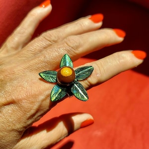 May include: A flower-shaped ring with green and blue enamel leaves and a brown center with a gold accent. The ring is worn on a finger with orange nail polish. The ring's design resembles a stylized flower.