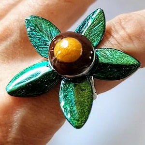 May include: A flower-shaped ring with green petals and a brown center with a yellow accent. The ring is made of metal and has a shiny, iridescent finish. The ring is worn on a finger.