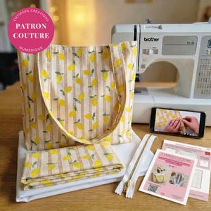 May include: A tote bag and a folded piece of fabric, both featuring a yellow lemon pattern on a cream background with thin brown vertical stripes. A white sewing machine, a smartphone displaying a sewing tutorial, and a printed instruction sheet.