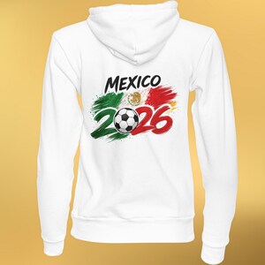 May include: White hooded sweatshirt featuring the word "MEXICO" in black, with a soccer ball and the number "2026" in red. The design incorporates the colors of the Mexican flag: green, white, and red, creating a patriotic theme.
