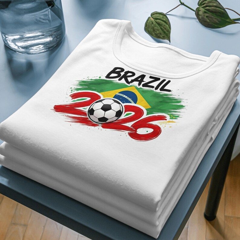 May include: A stack of white t-shirts with a Brazil-themed graphic. The design includes the word "BRAZIL" in black, the number "2026" in red, a soccer ball, and the Brazilian flag.