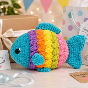 May include: A plush crochet fish toy featuring a vibrant rainbow color pattern with stripes of teal, purple, orange, yellow, lime green, and pink. The fish has a rounded body, a small fin, a tail, and black plastic eyes. It is made of soft yarn.