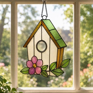 Op de afbeelding: Een glas-in-lood vogelhuisje zonnevanger met een witte behuizing, een groen dak en een geel zijpaneel. Een roze bloem met een geel hart en groene bladeren is aan de voorzijde bevestigd. Het hangt aan een zwarte metalen ketting met een zuignap.