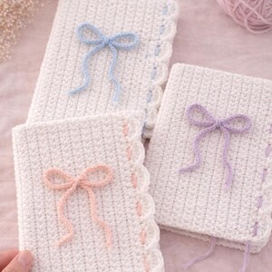 May include: Three white crocheted book covers with decorative bows in pastel colors: blue, pink, and purple. Each cover has a scalloped edge with matching thread accents. Yarn balls in pastel colors are in the background.