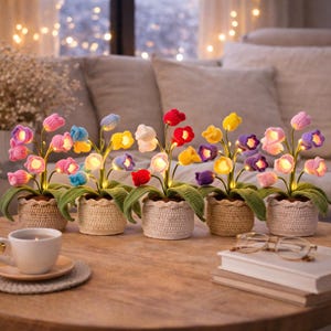 May include: A display of crochet flower lights in a range of colours, such as pink, blue, yellow, and purple, each in a small woven pot. The flowers are lit, producing a warm glow. The arrangement is on a wooden table.