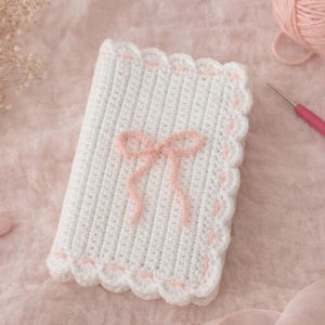 May include: A white crocheted blanket with a pink bow and scalloped edges. A pink crochet hook and a ball of pink yarn are also visible. The blanket is on a pink surface.