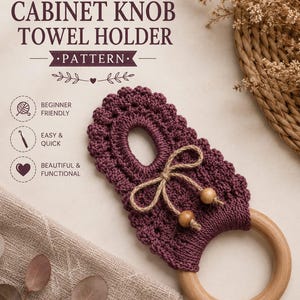 May include: A crochet cabinet knob towel holder pattern featuring a dark plum-colored crocheted piece with a scalloped edge, a central opening, and a natural wooden ring. The design includes a jute twine bow with two wooden beads attached.