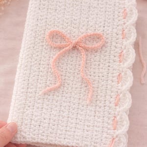 May include: A handmade, white crochet notebook cover with a delicate pink bow and scalloped edge. The cover is crafted with a tight stitch, and the bow adds a touch of elegance. The pink accents complement the neutral white, creating a soft, feminine aesthetic.