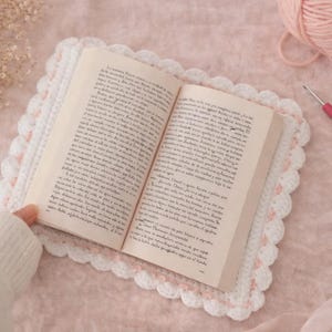 May include: An open book rests on a white and pink crocheted placemat. A pink crochet hook and a ball of pink yarn are next to the book. The scene is set on a pink surface, with a sprig of dried flowers and two pink heart-shaped candies.