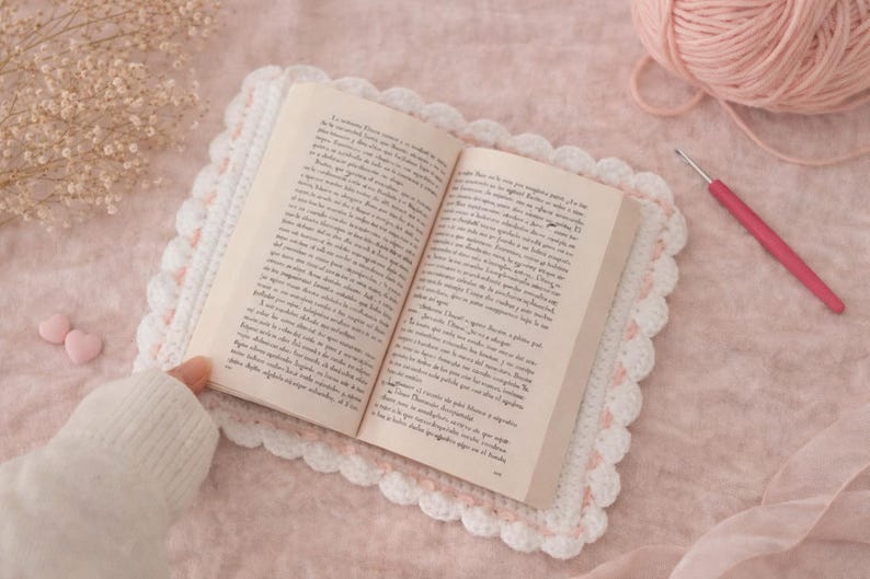 May include: An open book rests on a white and pink crocheted placemat. A pink crochet hook and a ball of pink yarn are next to the book. The scene is set on a pink surface, with a sprig of dried flowers and two pink heart-shaped candies.