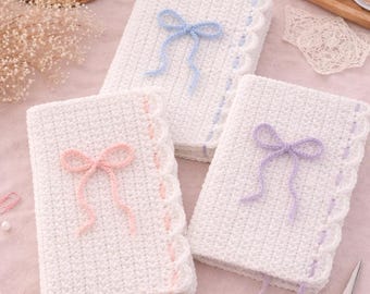 May include: Three white crochet notebooks with scalloped edges and yarn bows in blue, pink, and purple. The notebooks are on a pink surface with yarn, a cup of coffee, and scissors.