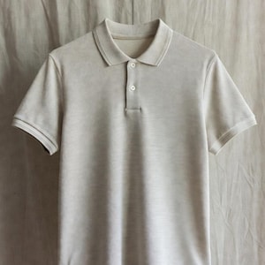 May include: A light beige polo shirt with short sleeves, a collar, and a three-button placket. The shirt is made of a textured fabric and hangs against a neutral-toned backdrop.