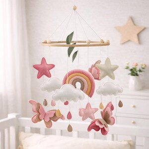 May include: A baby mobile featuring a wooden ring with hanging felt shapes. The mobile includes pink stars, white clouds, a rainbow, and butterflies in shades of pink and brown. Decorative leaves and teardrop accents complete the design.
