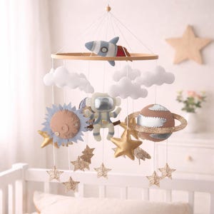 Op de afbeelding: Een baby mobile met een houten ring met een raket, wolken, een astronaut, een zon, een planeet met een glitterring en gouden sterren. De mobile is gemaakt van zachte materialen in blauw, bruin en goud.